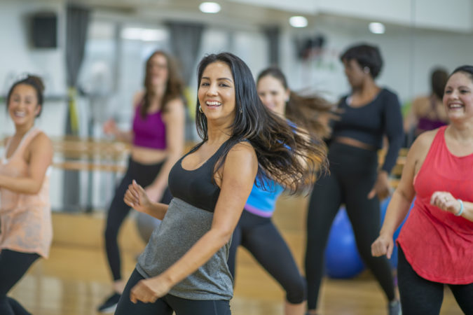 A multi-ethnic group of adult women are dancing in a fitness studio. They are wearing athletic clothes.
