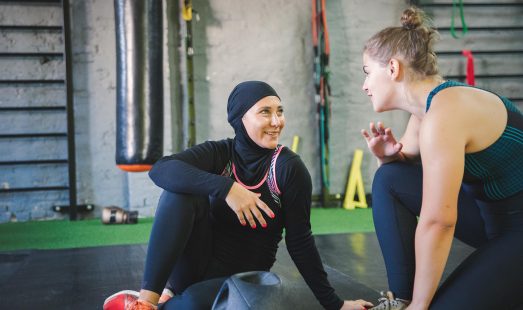 Two young females sat facing each other on the gym floor talking before or after a workout. Gym equuipment is located around them.