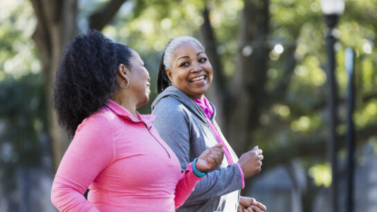 Two women exercising together in the city, jogging or power walking, laughing and conversing. Buildings and trees are out of focus in the background. The one in pink is in her 60s and her friend is in her 50s.