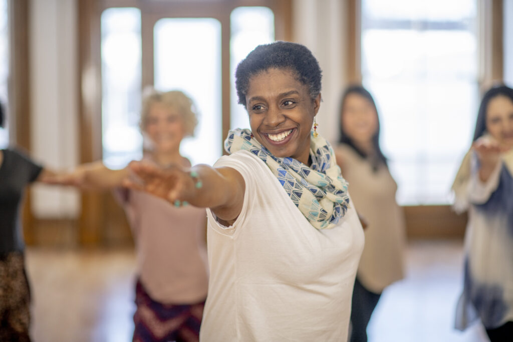 A senior female adult of African descent is participating in a group yoga class. The group is doing the Warrior Pose in class. She is happy and smiling.